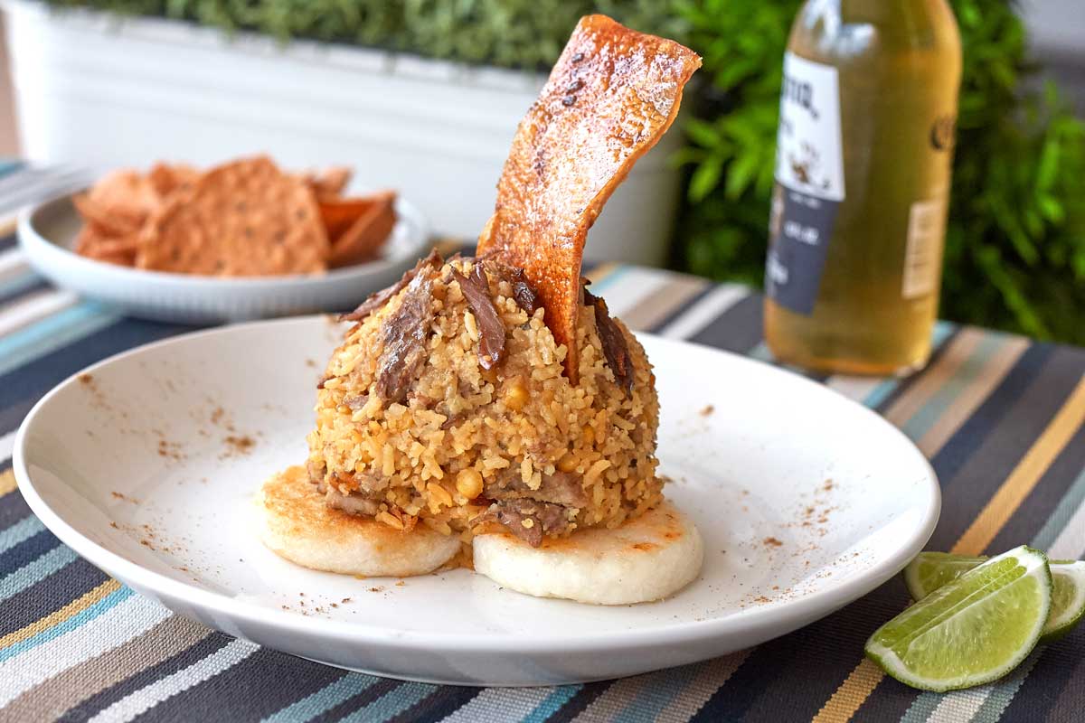 Gourmet Colombian lechona served with rice, shredded pork, and crispy pork skin on top of arepas, accompanied by lime wedges and a beer in the background.