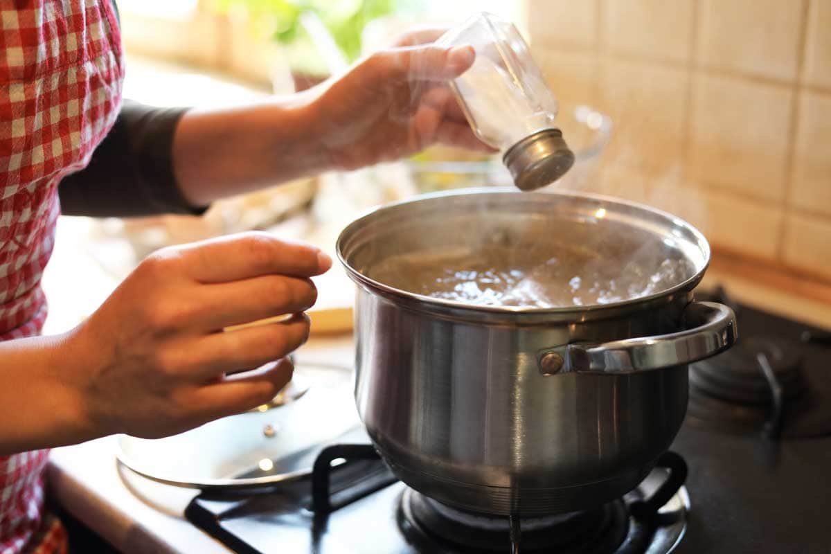 Close-up of a person adding salt to a steaming pot on the stove, preparing a homemade dish, possibly Caldo Gallego, in a cozy kitchen setting.