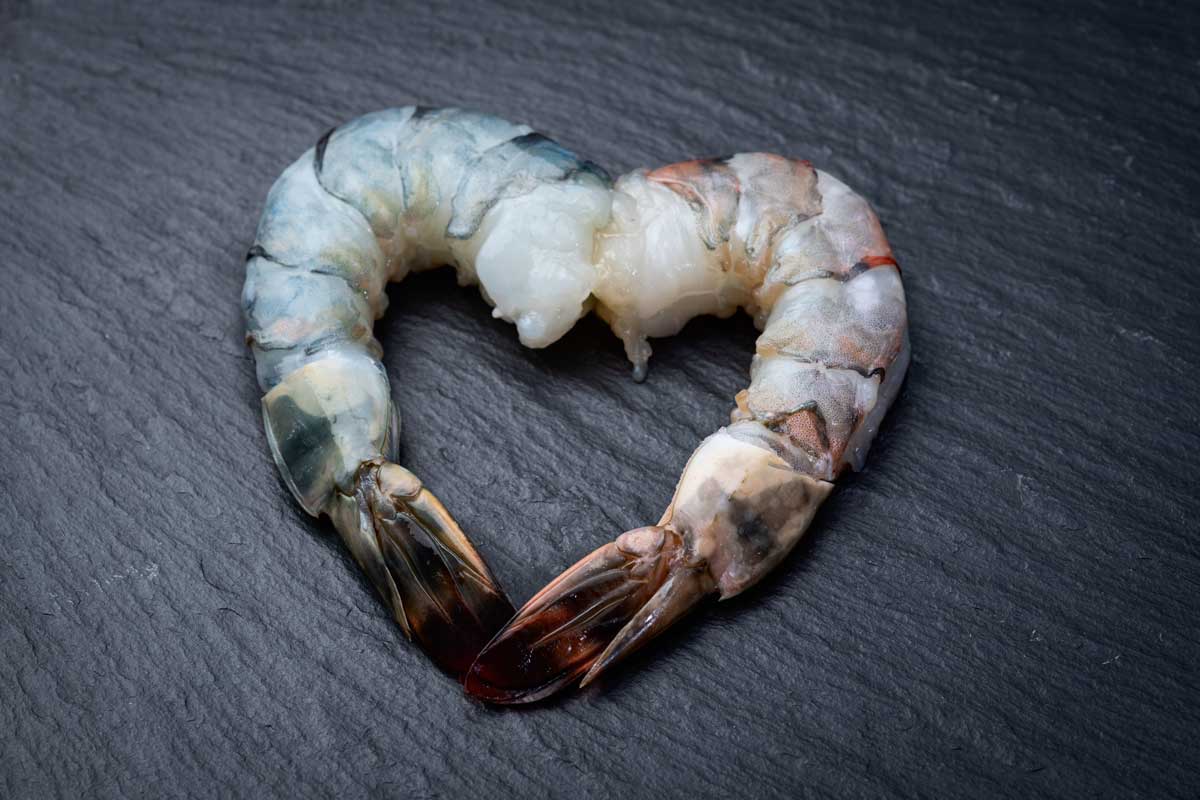 Two raw shrimp arranged in the shape of a heart on a dark slate surface, symbolizing the health benefits of seafood in Peruvian parihuela.