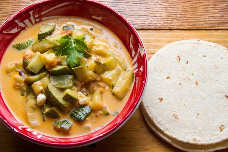 Bowl of creamy Mexican calabacitas with zucchini, corn, tomatoes, and cheese, garnished with cilantro, served alongside a stack of corn tortillas on a wooden table.