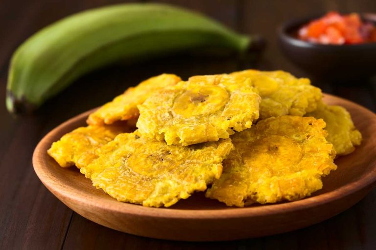Crispy fried patacones stacked on a wooden plate with a green plantain and salsa in the background.