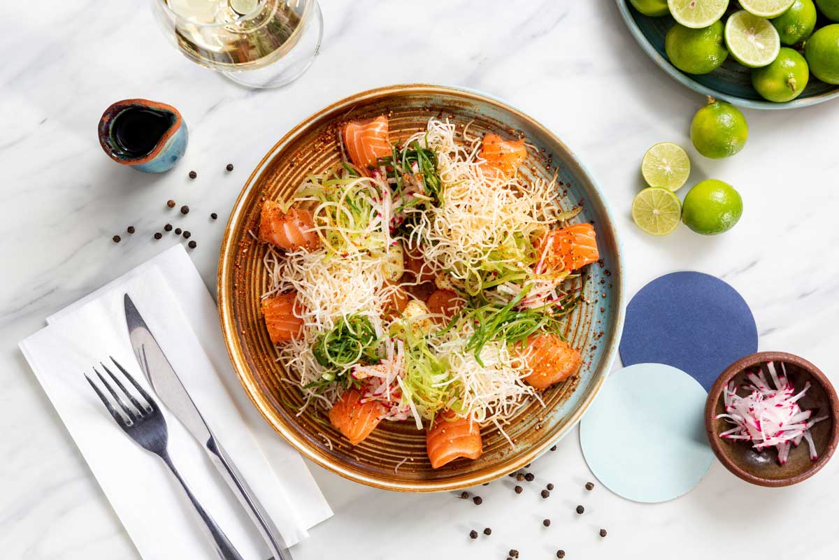 A beautifully plated Nikkei dish featuring fresh salmon slices, crispy noodles, shredded radish, and green garnishes on a ceramic plate, surrounded by limes, soy sauce, and black pepper on a marble surface.