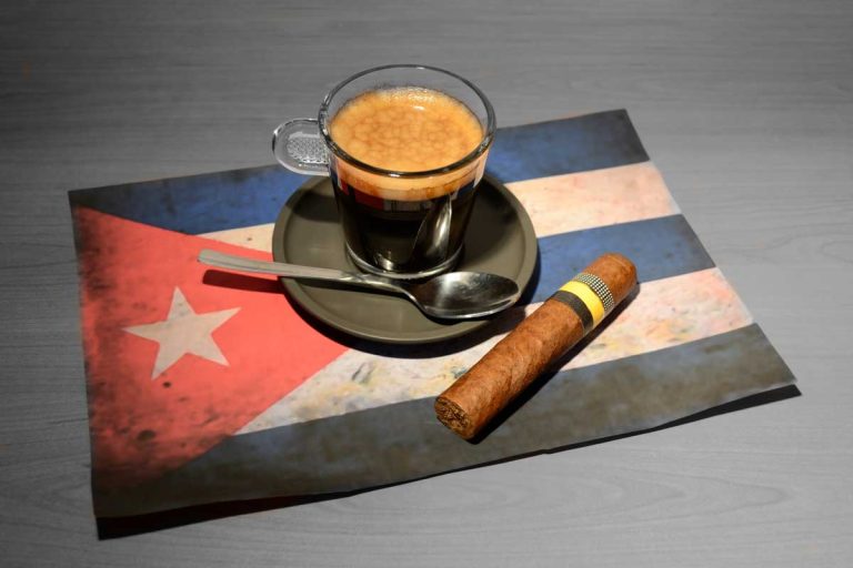 Glass cup of Cuban coffee with crema, a spoon, and a Cuban cigar placed on a Cuban flag-themed placemat on a wooden table.