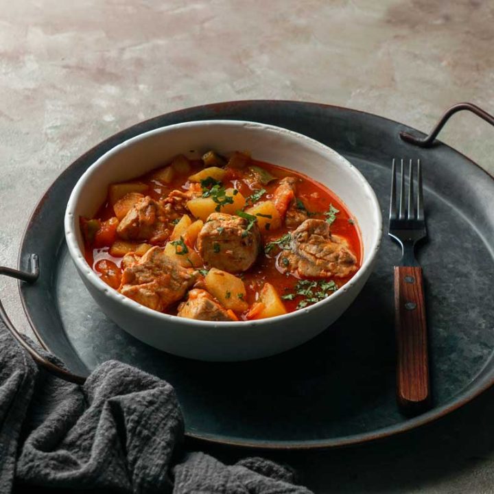 A rustic bowl of Marmitako, the traditional Basque tuna and potato stew, with chunks of tuna, potatoes, and peppers in a rich red broth, garnished with fresh herbs and served on a round metal tray with a fork.