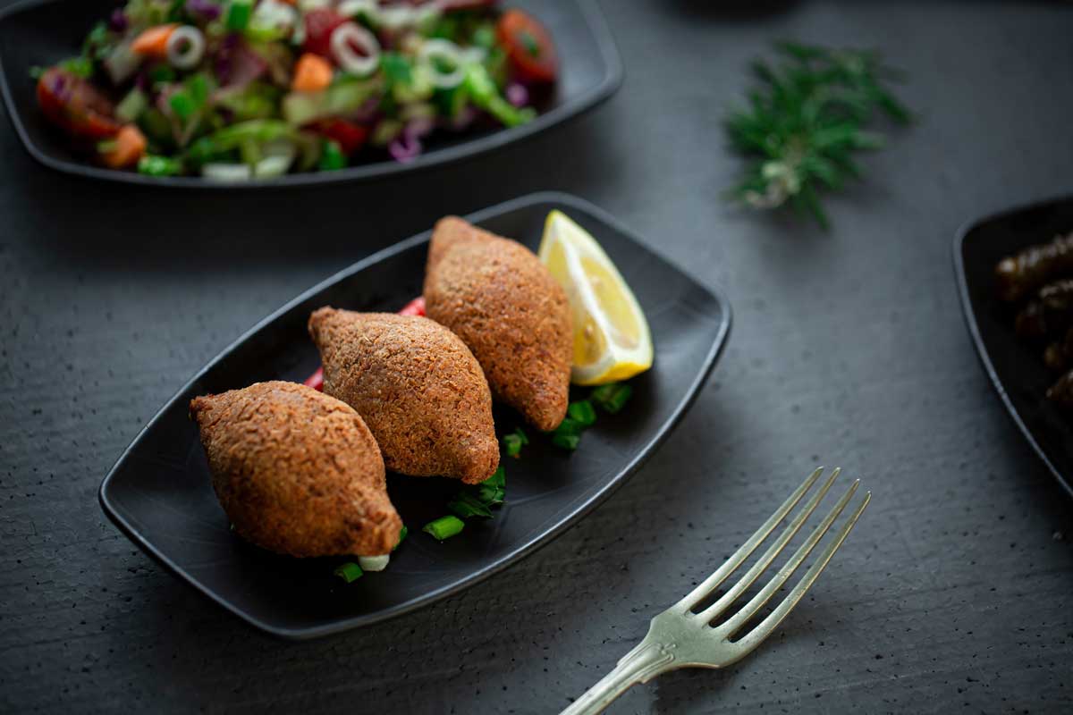 Three golden-brown Dominican kipes served on a black plate with lemon wedges and chopped green onions, with a salad and fork visible in the background.